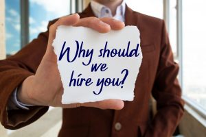 Closeup On Businessman Holding A Card With WHY SHOULD WE HIRE YO